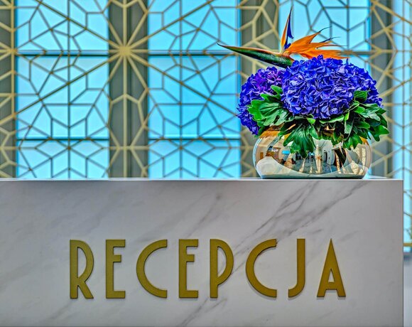Marmurowa recepcja ze złotymi literami „RECEPCJA”, bukietem niebieskich hortensji i ptakiem rajskim w lustrzanym wazonie.