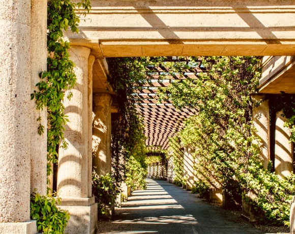 Hotelowa pergola z kamiennymi kolumnami i bujnymi pnączami, korytarz ogrodowy z cieniem i słońcem, sprzyja relaksującym spacerom.
