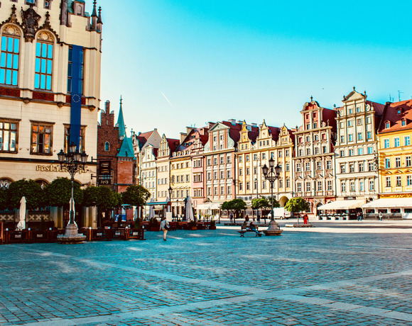 Sunny historic town square with colorful baroque facades, cobblestone plaza, streetlamps and outdoor restaurant terraces.
