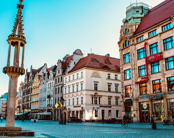 Zabytkowy rynek z ozdobną kolumną, brukowanym placem, kolorowymi kamienicami i kawiarniami z tarasami.