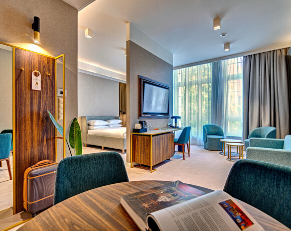 Spacious modern hotel suite with king bed, seating area and teal chairs, work desk, TV, large sunlit window.