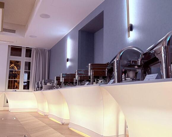 Beleuchtetes Buffet mit glänzenden Wärmeschalen, Tellerstapel und eleganter Veranstaltungslounge vor großen Fenstern.