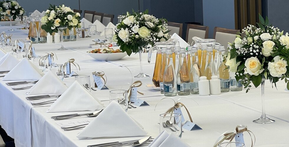 Długi stół bankietowy z białym obrusem, złożonymi serwetkami, srebrnymi sztućcami, butelkami i bukietami białych róż