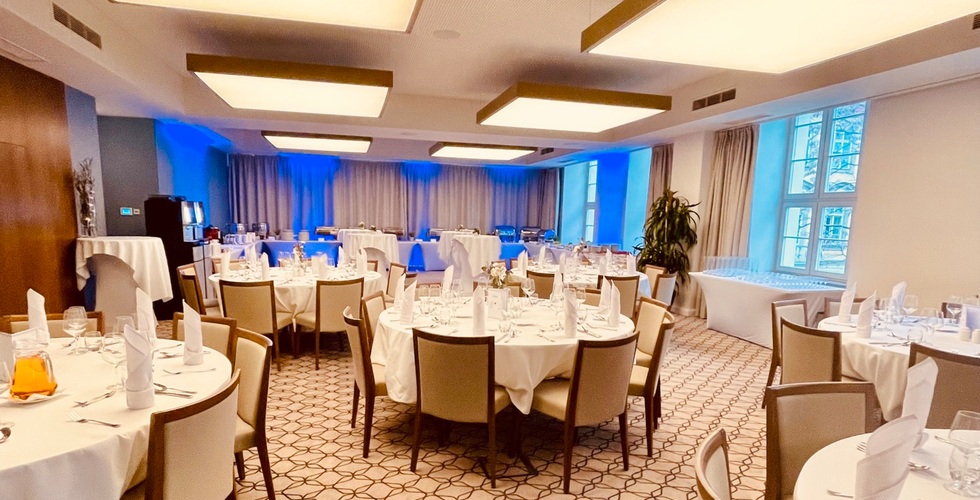 Elegancka sala bankietowa z okrągłymi stołami, białymi obrusami, złożonymi serwetkami, bufetem i niebieskim oświetleniem.