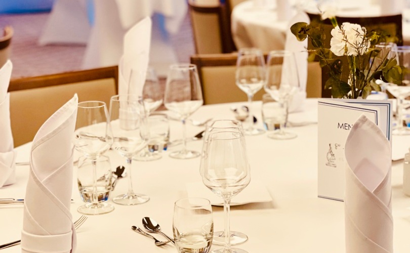 Elegancko nakryty stół bankietowy z białym obrusem, złożonymi serwetkami, kieliszkami, sztućcami, kartą menu i kwiatowym centrum pod niebieskim światłem