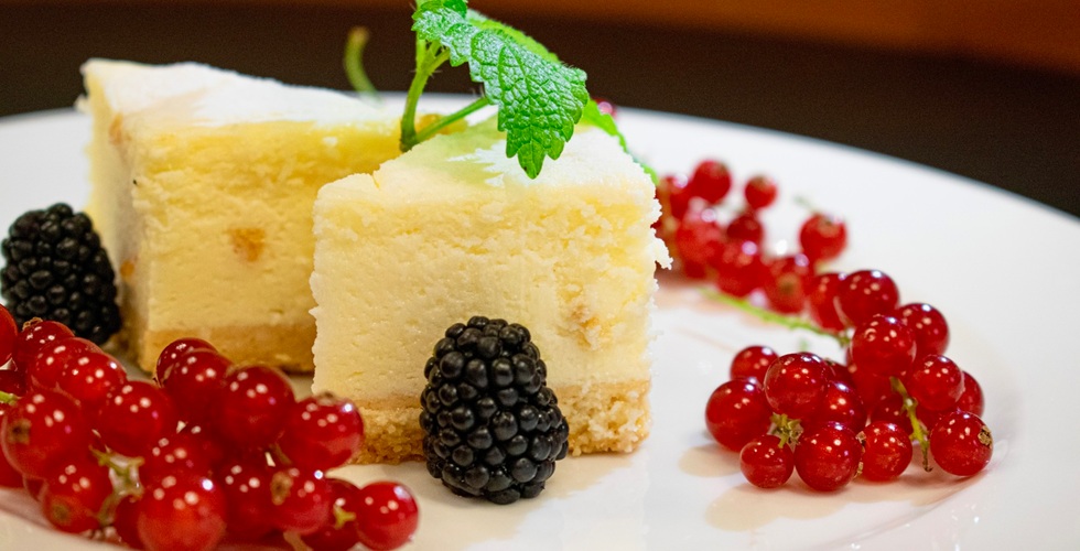 Kawałki mini sernika na maślanym spodzie, udekorowane czerwonymi porzeczkami, jeżyną i świeżą miętą na białym talerzu