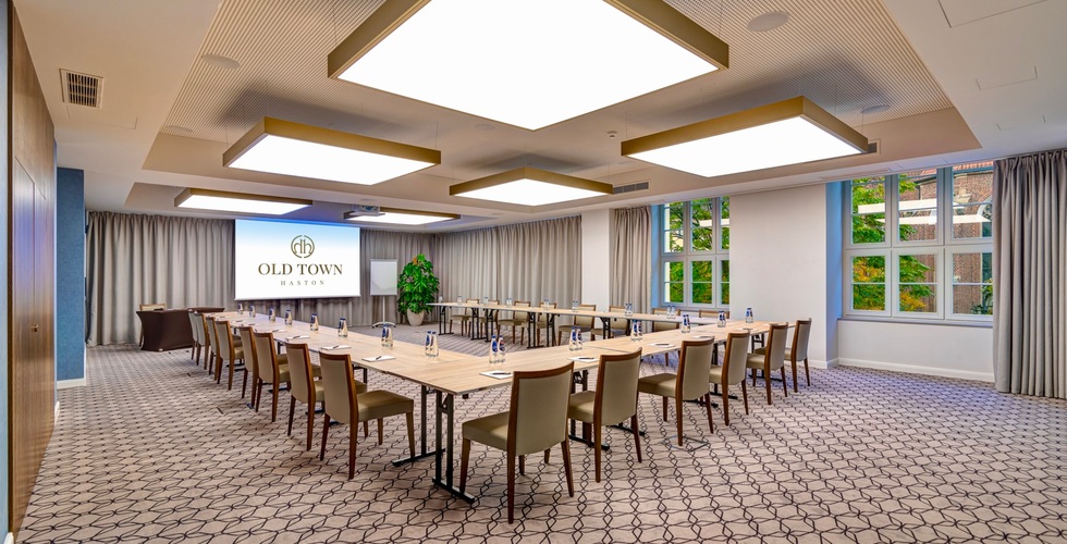 Nowoczesna sala konferencyjna w układzie U: drewniane stoły, tapicerowane krzesła, butelki wody, ekran z logo Old Town Haston, duże kwadratowe lampy