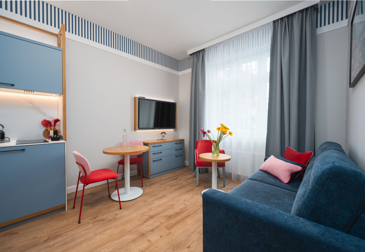 Nowoczesny pokój hotelowy z aneksem kuchennym, niebieską sofą, czerwonymi krzesłami i dwoma stolikami z kwiatami