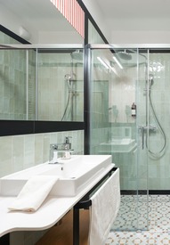 Hotelowa łazienka z przeszklonym prysznicem z deszczownicą, zielonymi płytkami, wzorzystą podłogą, umywalką z ręcznikiem, paskowaną ścianą i pralką.