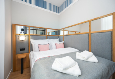 Przytulny pokój hotelowy z miękkim łóżkiem, szarym zagłówkiem, różowymi poduszkami i ciepłym oświetleniem ściennym