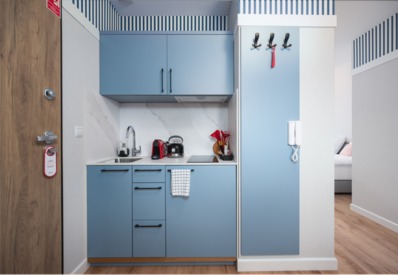 Kompaktowa hotelowa aneks kuchenny z niebieskimi szafkami, czajnikiem, ekspresem do kawy i przytulnym wejściem do pokoju.