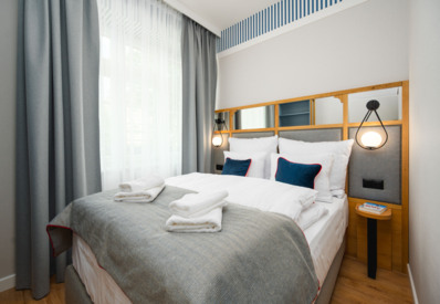 Przytulny pokój hotelowy z podwójnym łóżkiem, szarym narzutem, granatowymi poduszkami i miękkim oświetleniem.