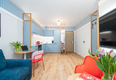 Jasne studio hotelowe z niebieską kuchnią, ekspresem do kawy, biurkiem z laptopem, czerwonymi fotelami, tulipanami i telewizorem.