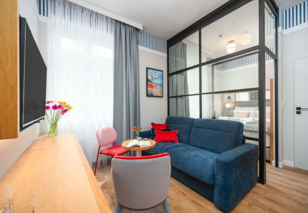 Nowoczesny pokój hotelowy z niebieską sofą, czerwonymi i różowymi krzesłami, oddzieloną sypialnią ze szklaną przegrodą