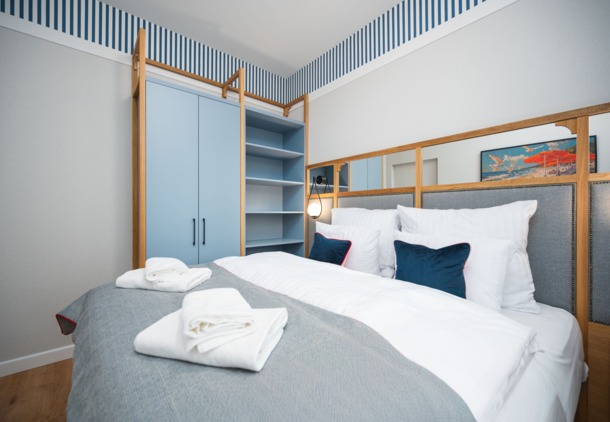 Przytulny pokój z wygodnym łóżkiem, białą pościelą i granatowymi poduszkami, niebieską szafą i paskami na ścianie