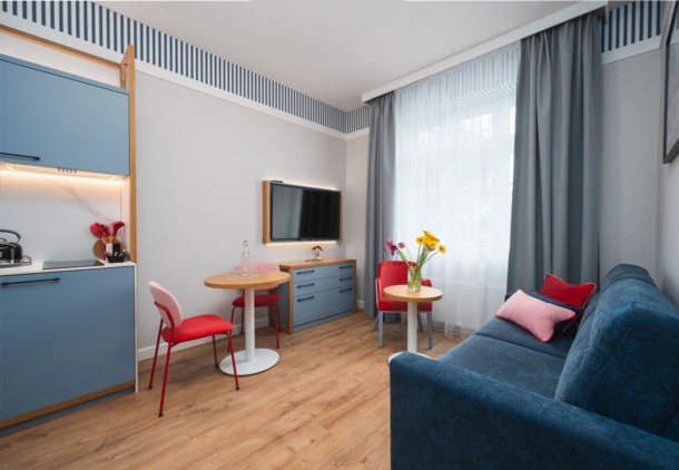 Nowoczesny pokój hotelowy z aneksem kuchennym, niebieską sofą, czerwonymi krzesłami i dwoma stolikami z kwiatami