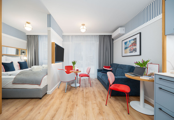 Jasne, nowoczesne studio z łóżkiem, salonem z granatową sofą, stolikiem jadalnym, biurkiem, telewizorem na ścianie i czerwonymi krzesłami