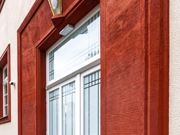 Wejście do hotelu z białymi, zdobionymi drzwiami i rustykalną, czerwoną oprawą z lampą nad drzwiami