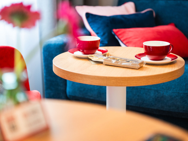 Dwie czerwone filiżanki na drewnianym stoliku, wygodna kanapa z niebieskimi i czerwonymi poduszkami w tle