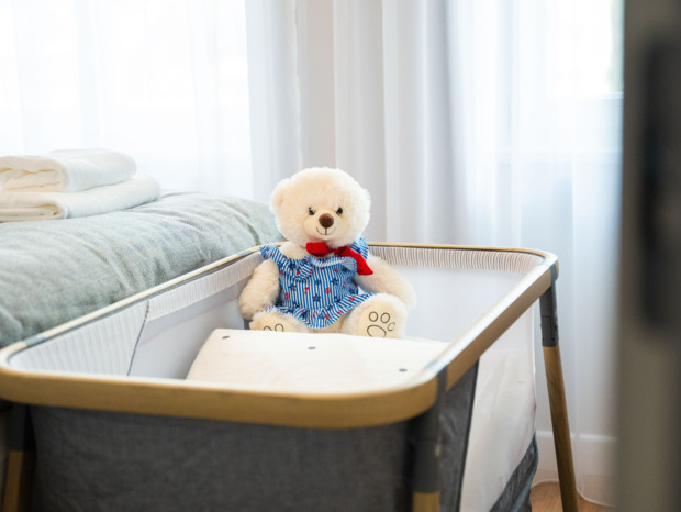 Przytulne łóżeczko niemowlęce z pluszowym misiem w sukience, obok łóżko hotelowe z czystymi ręcznikami.