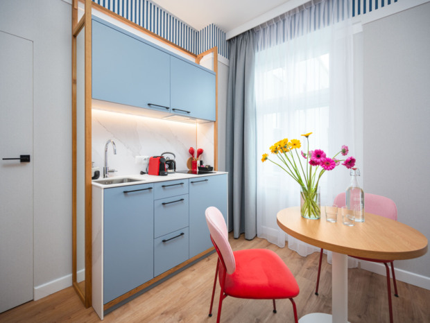 Kompaktowa kuchnia z niebieskimi szafkami, stolem z kwiatami i czerwonymi krzesłami w jasnym, nowoczesnym pokoju hotelowym.