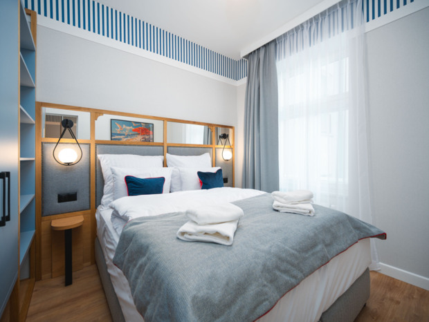 Przytulny pokój hotelowy z podwójnym łóżkiem w niebieskich i białych odcieniach, dużym oknem i nowoczesnym oświetleniem.