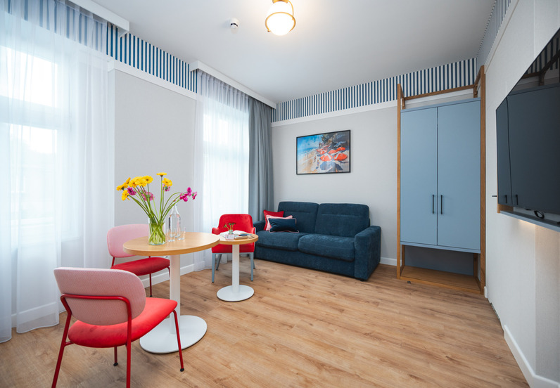 Jasny hotelowy salon z niebieską sofą, czerwonymi krzesłami, okrągłym stolikiem z kwiatami, szafą, telewizorem i przezroczystymi zasłonami