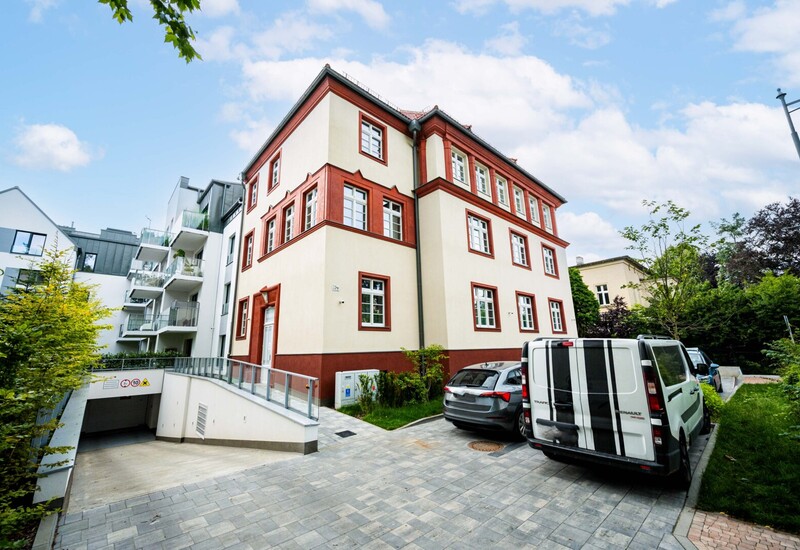 Trzykondygnacyjny narożny budynek w kremie z czerwonymi ramami okien, brukowany podjazd z zaparkowanymi samochodami, zjazd do garażu i balkony
