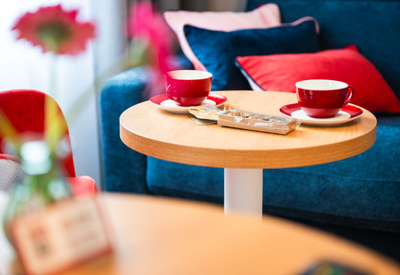 Dwie czerwone filiżanki na drewnianym stoliku, wygodna kanapa z niebieskimi i czerwonymi poduszkami w tle