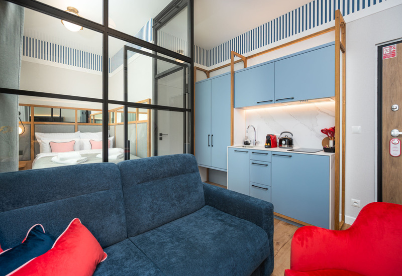 Studio hotelowe z granatową sofą, szklaną przegrodą do sypialni, niebieską kuchnią z ekspresem do kawy i marmurowym blatem