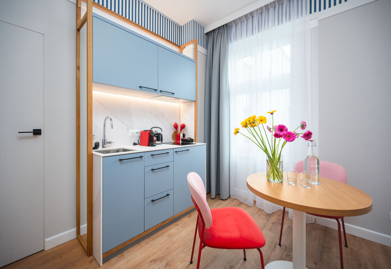 Kompaktowa kuchnia z niebieskimi szafkami, stolem z kwiatami i czerwonymi krzesłami w jasnym, nowoczesnym pokoju hotelowym.