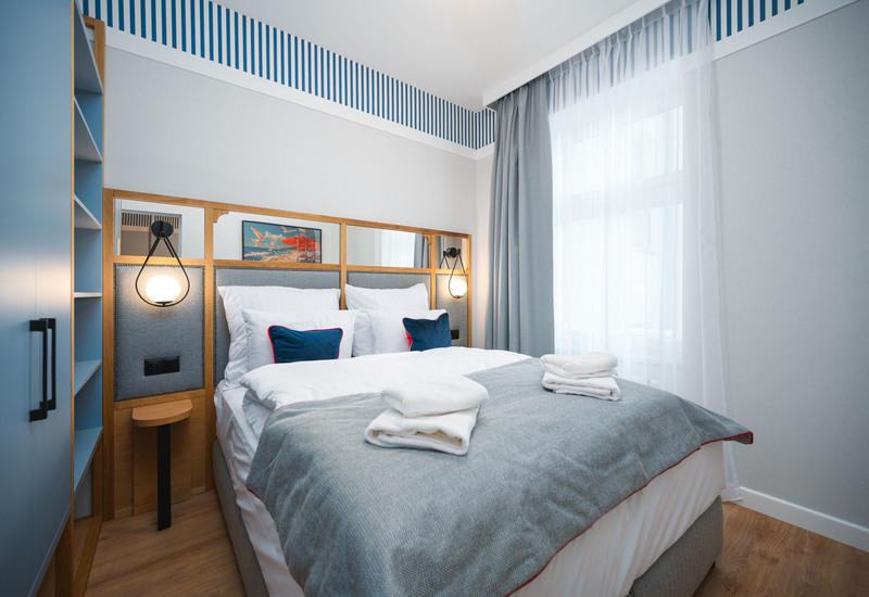 Przytulny pokój hotelowy z podwójnym łóżkiem w niebieskich i białych odcieniach, dużym oknem i nowoczesnym oświetleniem.