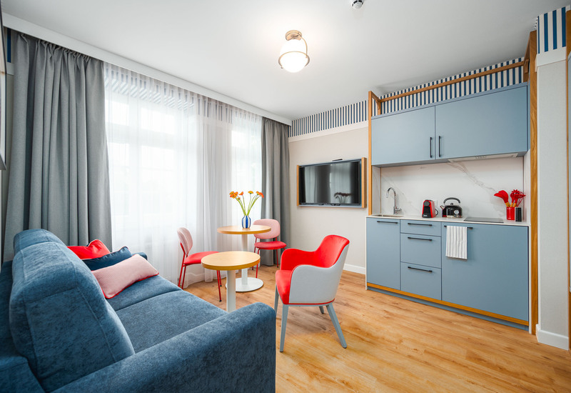 Jasne studio hotelowe z niebieską sofą, aneksem kuchennym z niebieskimi szafkami i ekspresem do kawy, stolikiem, czerwonymi krzesłami i telewizorem