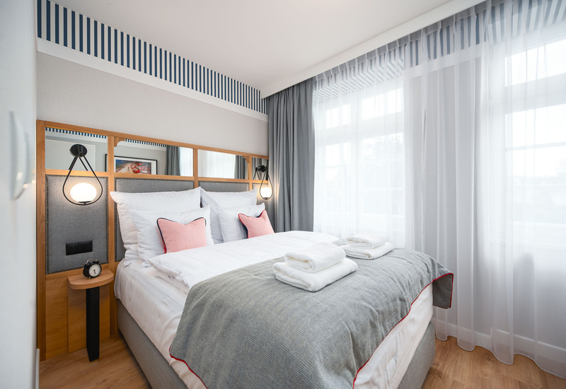 Jasny pokój dwuosobowy z wygodnym łóżkiem, białą pościelą, różowymi poduszkami, złożonymi ręcznikami, lampami przy łóżku i zwiewnymi zasłonami