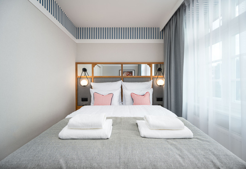 Jasny pokój hotelowy z łóżkiem dwuosobowym, białą pościelą, dwoma różowymi poduszkami, lampkami przy łóżku i złożonymi ręcznikami