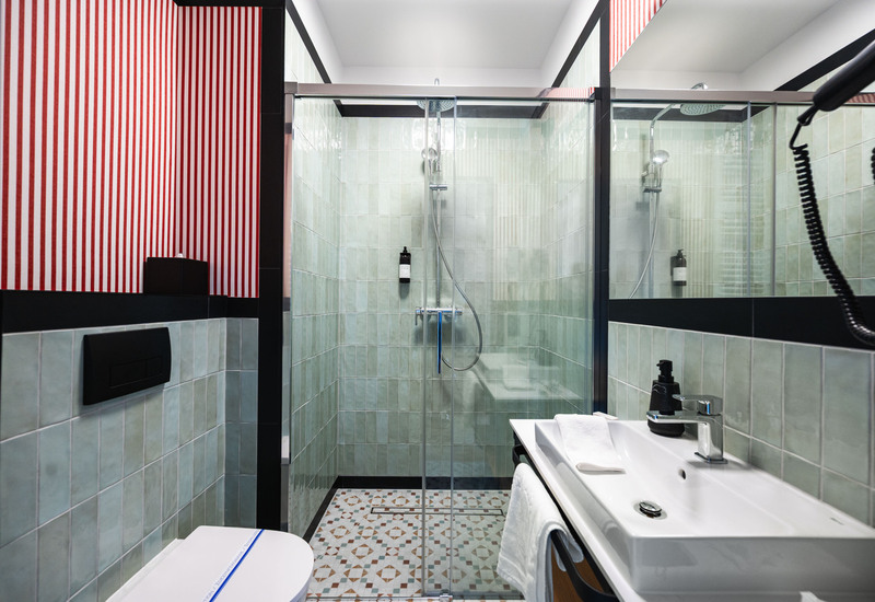 Nowoczesna łazienka hotelowa z przeszklonym prysznicem deszczowym, zielonymi płytkami, wzorzystą posadzką i umywalką z ręcznikiem.