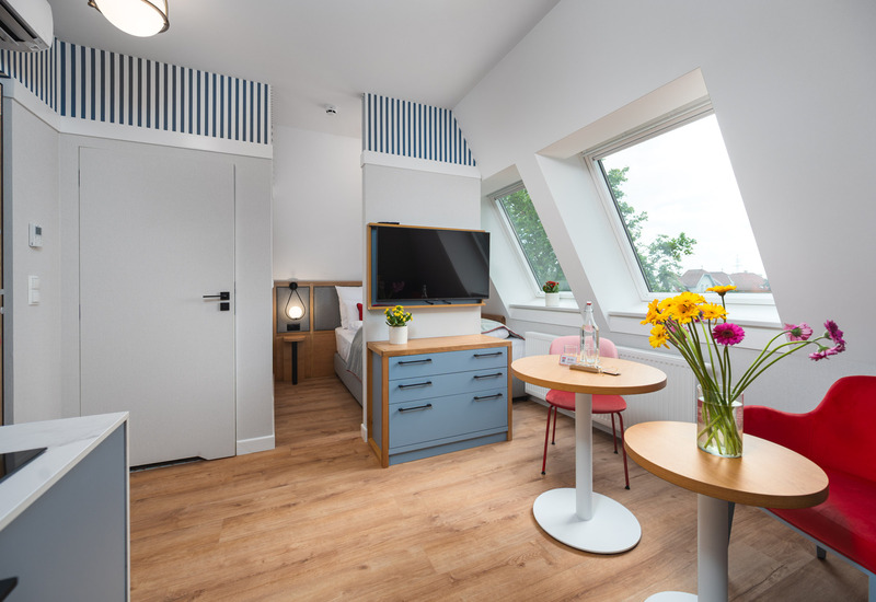 Jasny pokój na poddaszu z pochyłymi oknami dachowymi, łóżkiem, telewizorem nad niebieską komodą, stolikiem i kwiatami