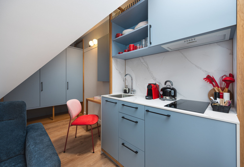 Kompaktowy aneks kuchenny w studio z niebieskimi szafkami, marmurowym panelem, ekspresem do kawy i czajnikiem, płytą indukcyjną i krzesłem