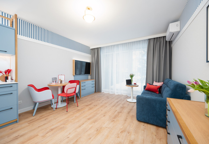 Jasne, nowoczesne studio z niebieską sofą, czerwonymi krzesłami, okrągłym stolikiem, TV, aneksem i dużym oknem z zasłonami