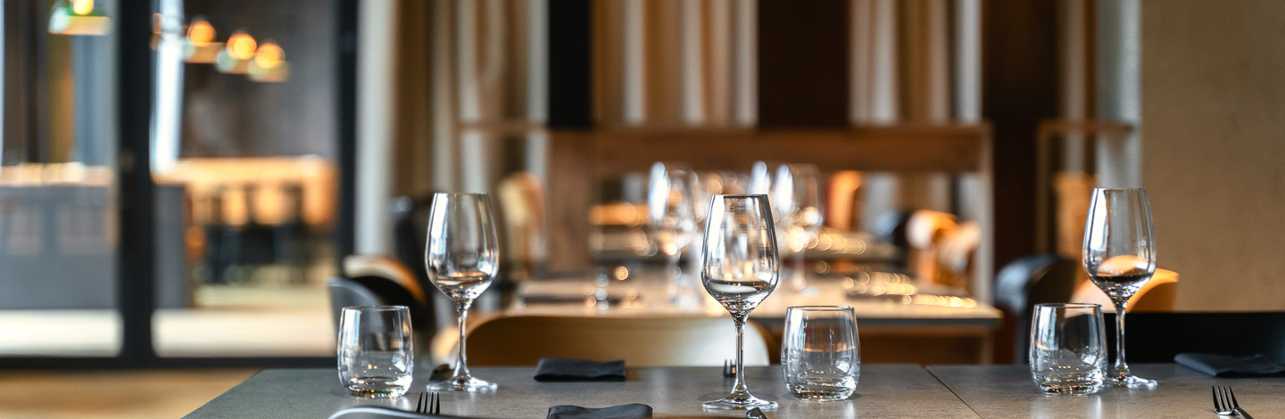 Elegancka restauracja z nowoczesnym wystrojem, stoliki nakryte kieliszkami i sztućcami, przytulna atmosfera.