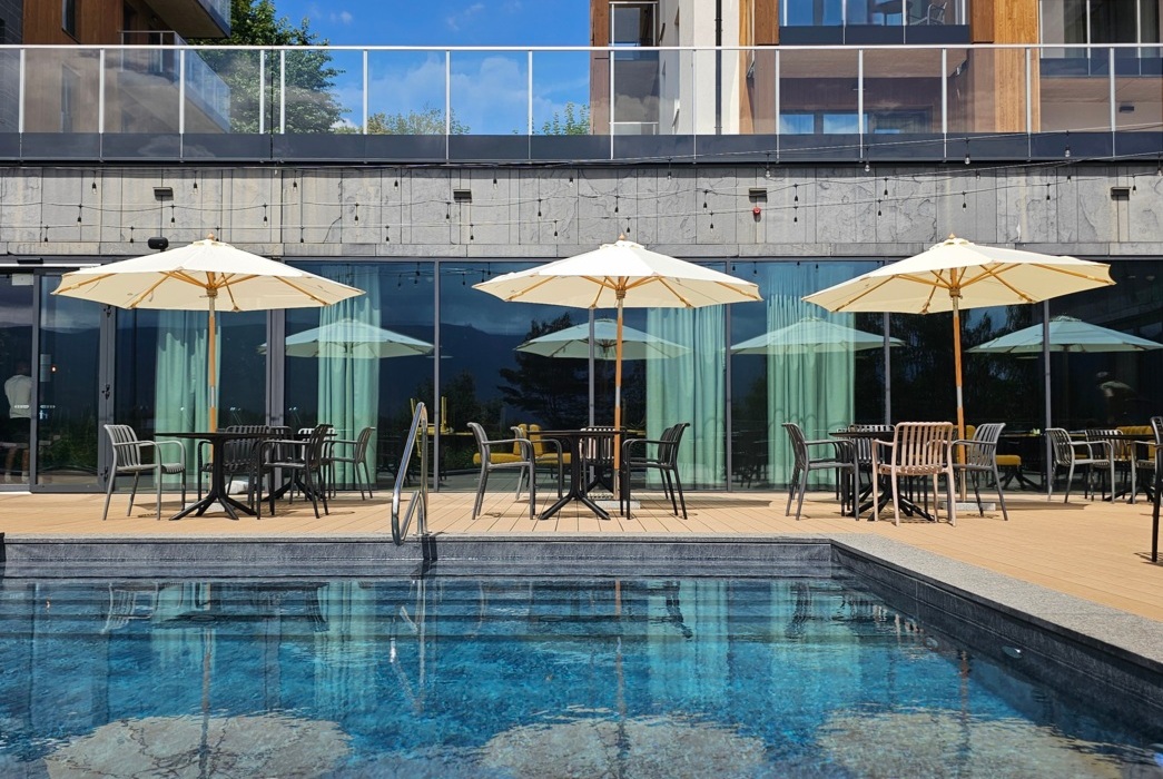 Taras przy basenie hotelowym z trzema stołami, krzesłami i parasolami na tle nowoczesnego budynku z drewnianymi akcentami.