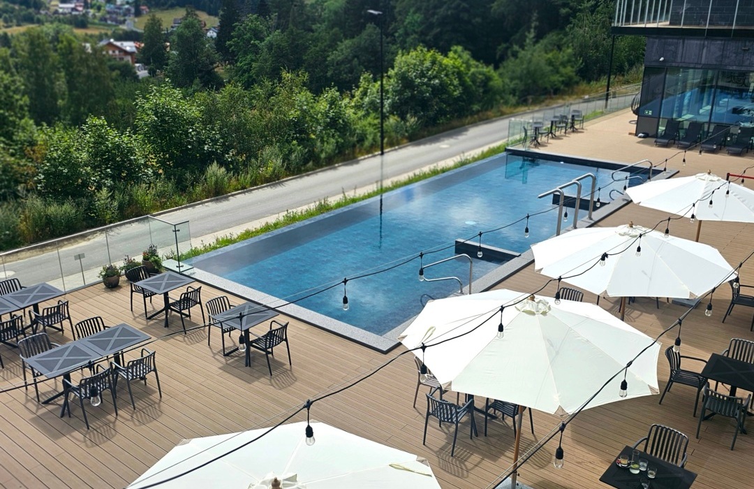 Taras hotelowy z basenem bez krawędzi, stolikami, parasolami i widokiem na zielone wzgórza i las.