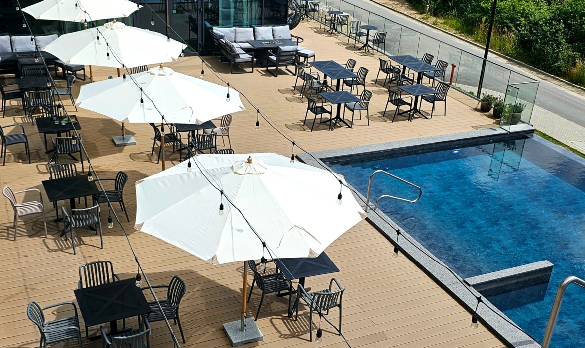 Taras hotelowy z odkrytym basenem, stolikami z czarnymi krzesłami i białymi parasolami dla gości.