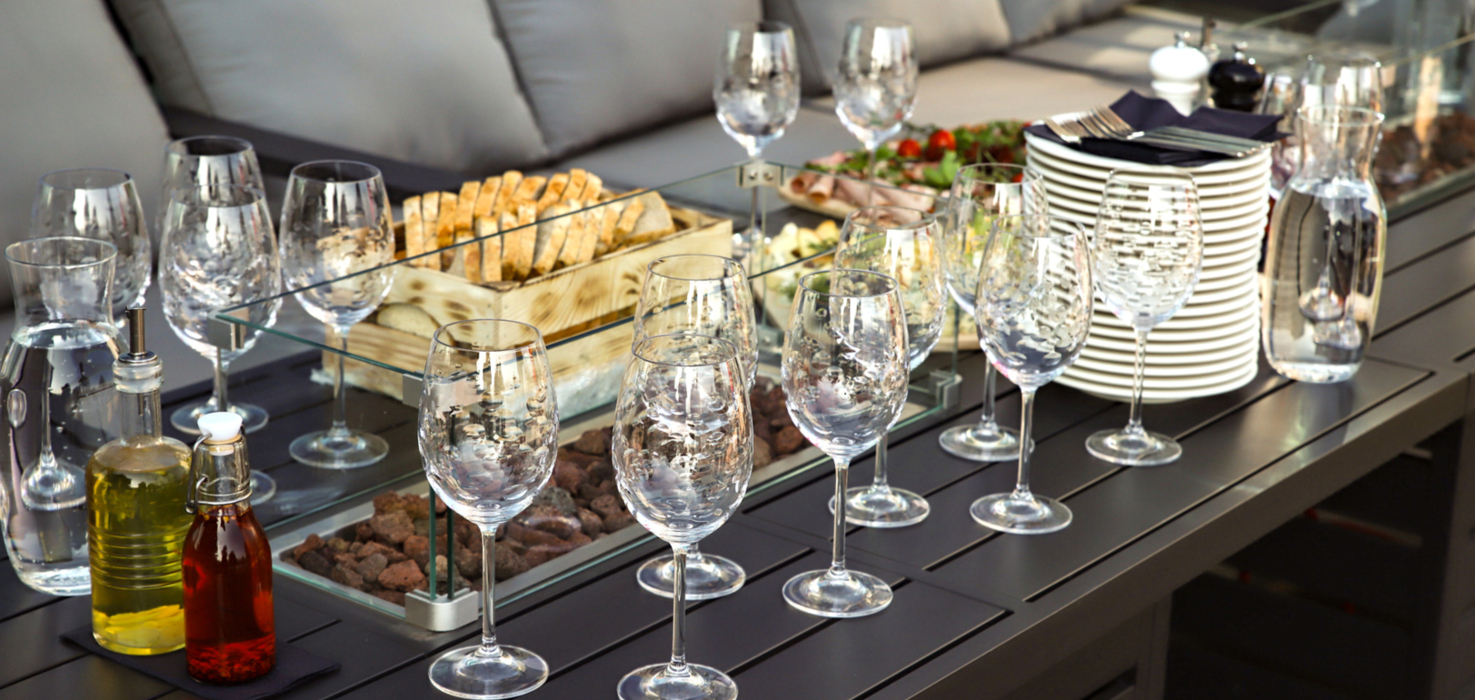 Elegancki stół z kryształowymi kieliszkami, talerzami i przystawkami, ustawiony obok szarej kanapy na tarasie.