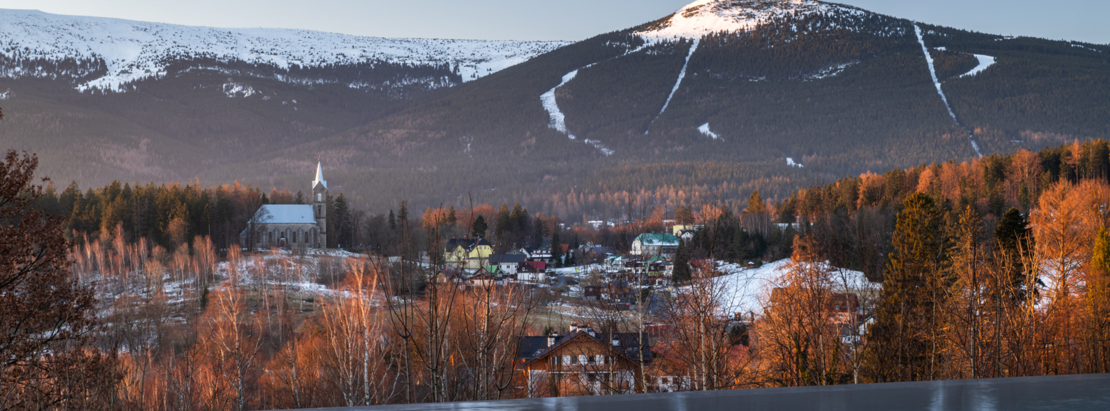 Infinity-Pool mit Blick auf schneebedeckte Berge, malerisches Dorf mit Kirche und herbstlichen Bäumen bei Dämmerung