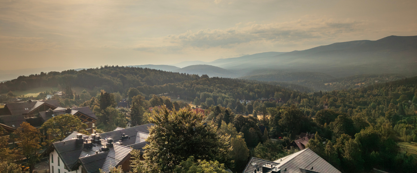 Horský resort s moderními domy mezi zelenými stromy a mlhavými kopci za teplého slunečního svitu.