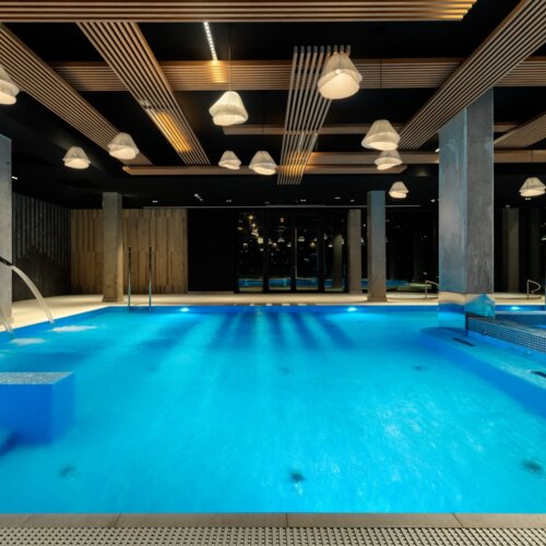 Moderner überdachter Hotelpool mit blauem Wasser, Wasserstrahlen, Deckenbeleuchtung und hölzernen Designelementen.