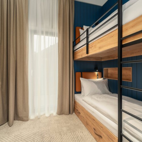 Modernes Hotelzimmer mit Etagenbett aus Holz, weicher weißer Bettwäsche und tiefblauer Akzentwand neben beigen Vorhängen