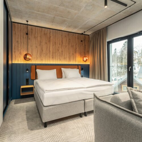 Modernes Hotelzimmer mit Doppelbett, Holzpaneelwand, hängenden Lampen und großem Fenster mit Blick auf bewaldete Hügel.