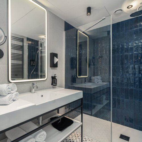 Moderne Hotelbadezimmer mit beleuchtetem Spiegel, weißem Waschbecken und begehbarer Dusche mit dunkelblauen Fliesen
